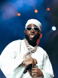 Afrobeats musician Davido performing live on stage wearing a white outfit, white sunglasses, and a diamond chain necklace.