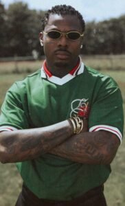 Asake wearing a green knit polo with red floral embroidery, gold oval sunglasses, and gold bracelets in the MCBH music video.