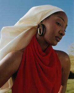Tems wearing a red Givenchy open-back silk top, a cream twisted silk-satin headpiece, and chunky black leather hoop earrings against a blue sky.