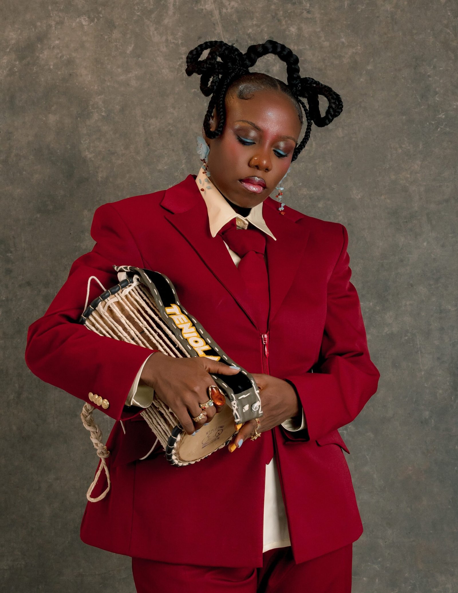 Teni holding a traditional Yoruba talking drum custom-marked with her name TENIOLA, dressed in a crimson double-breasted suit and matching red tie.