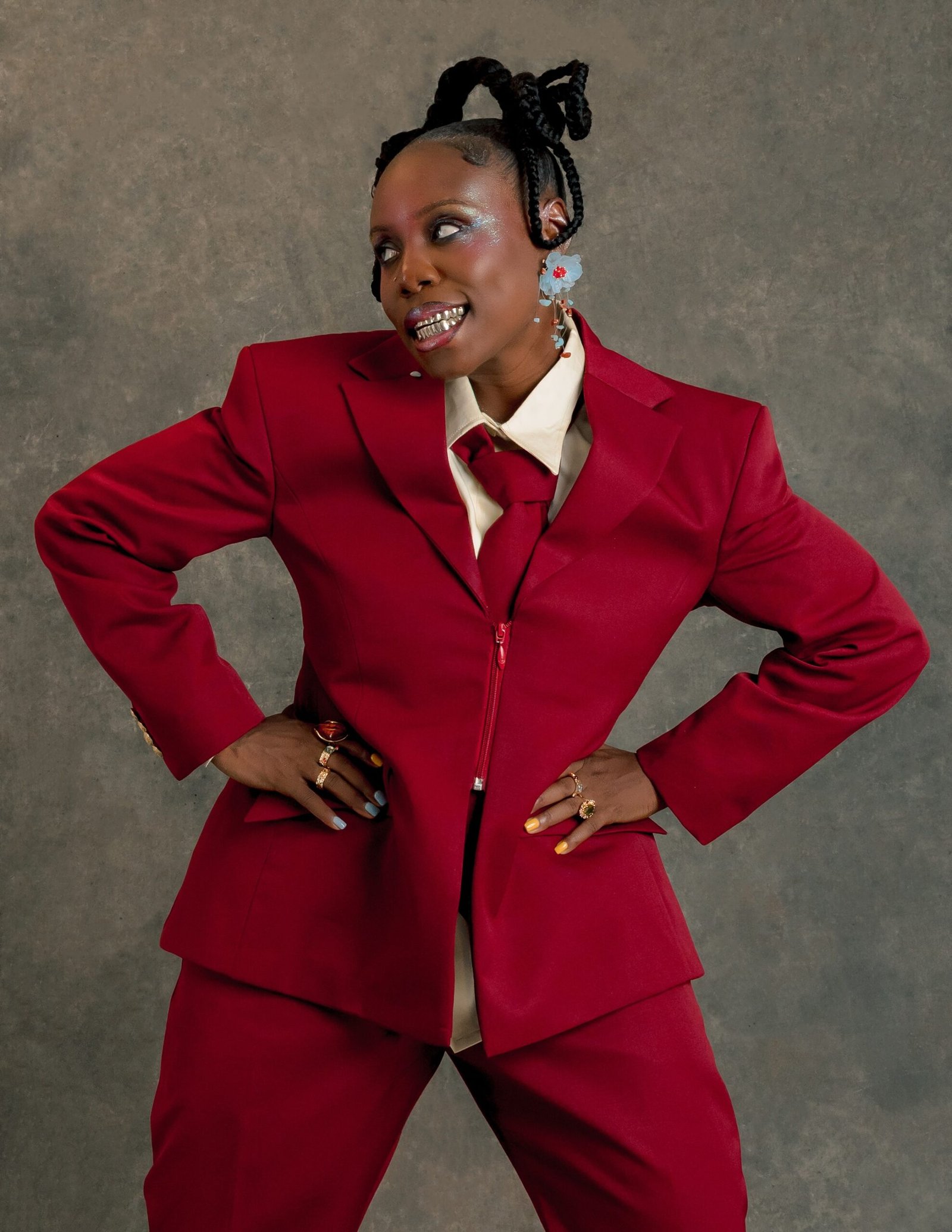 Teni striking a dynamic pose with hands on hips in an oversized crimson suit, featuring sculpted horn-shaped braids and blue eyeshadow.