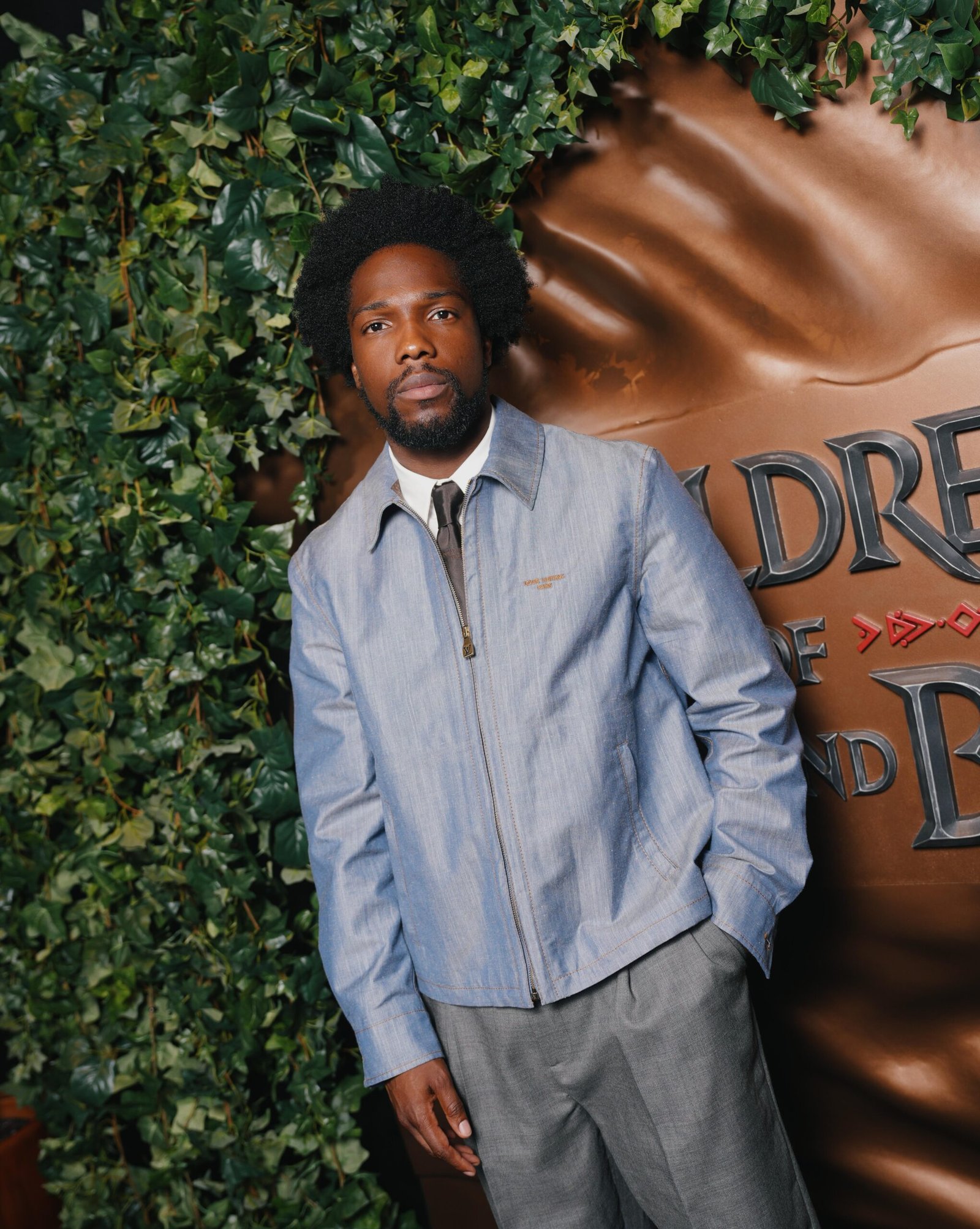 Actor Tosin Cole wearing a light blue denim shirt standing in front of the Children of Blood and Bone backdrop in Brooklyn.