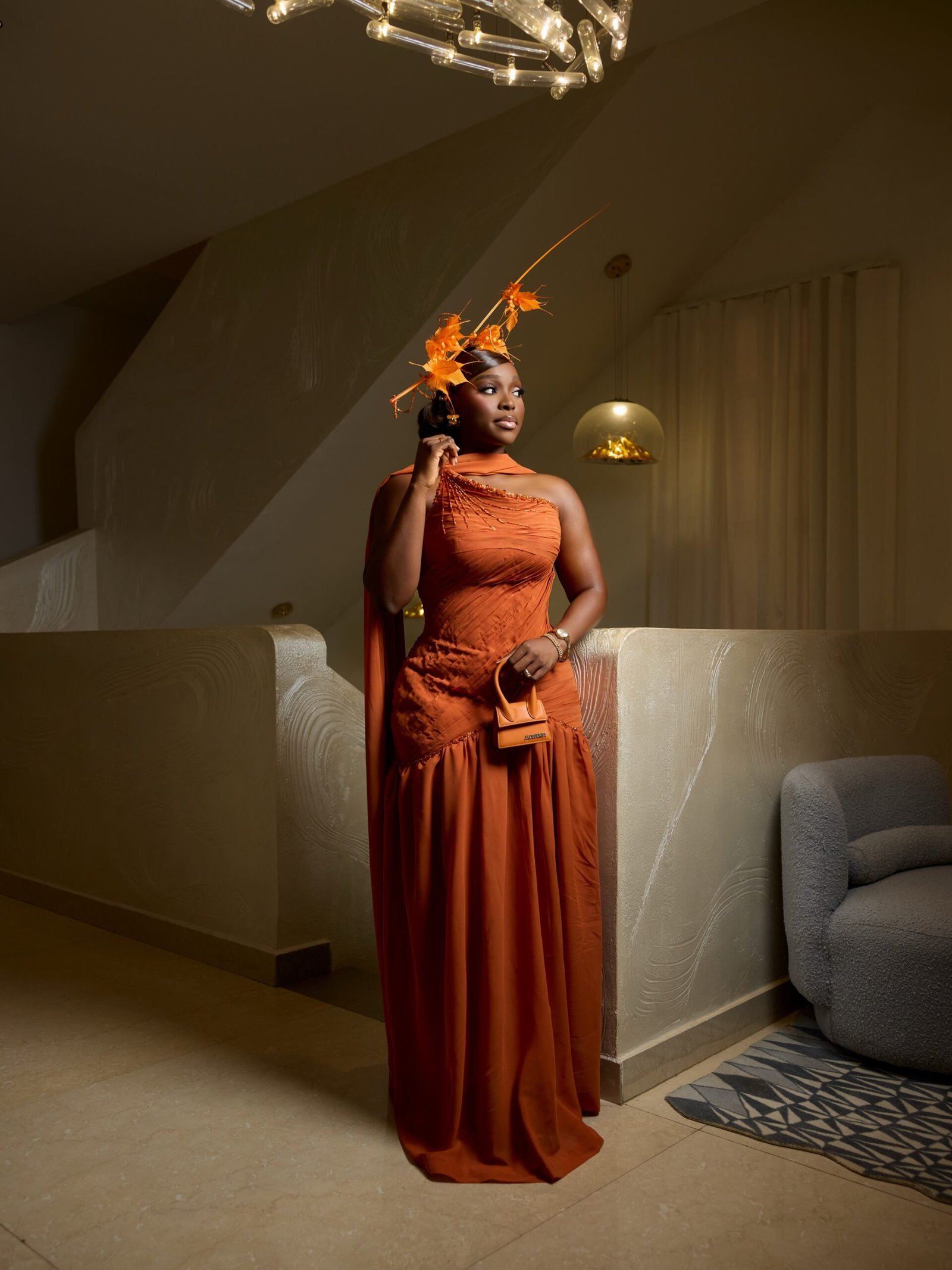 Tomike Adeoye in a burnt orange ruched asymmetric gown with beaded shoulder scarf, leaf-and-feather fascinator, and a matching orange Jacquemus mini bag.