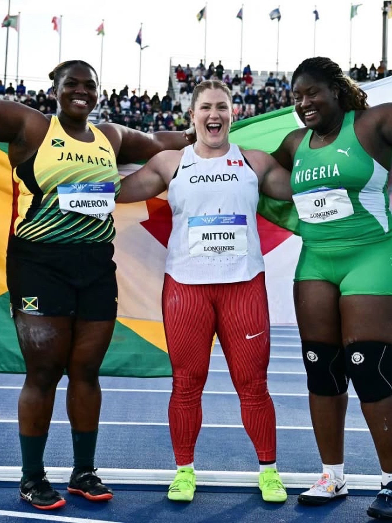 Commonwealth Games 2026 women's shot put medallists Lloydricia Cameron of Jamaica, Sarah Mitton of Canada, and Jessica Oji of Nigeria celebrating together on the track with national flags.