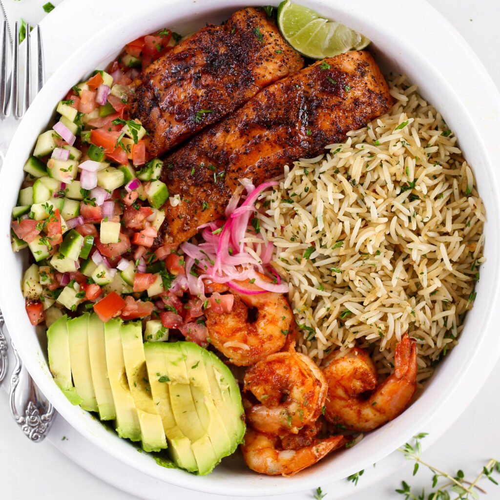 Kikifoodies garlic herb rice served in a bowl with pan-seared salmon, grilled shrimp, sliced avocado, pickled onions, and cucumber tomato salad.