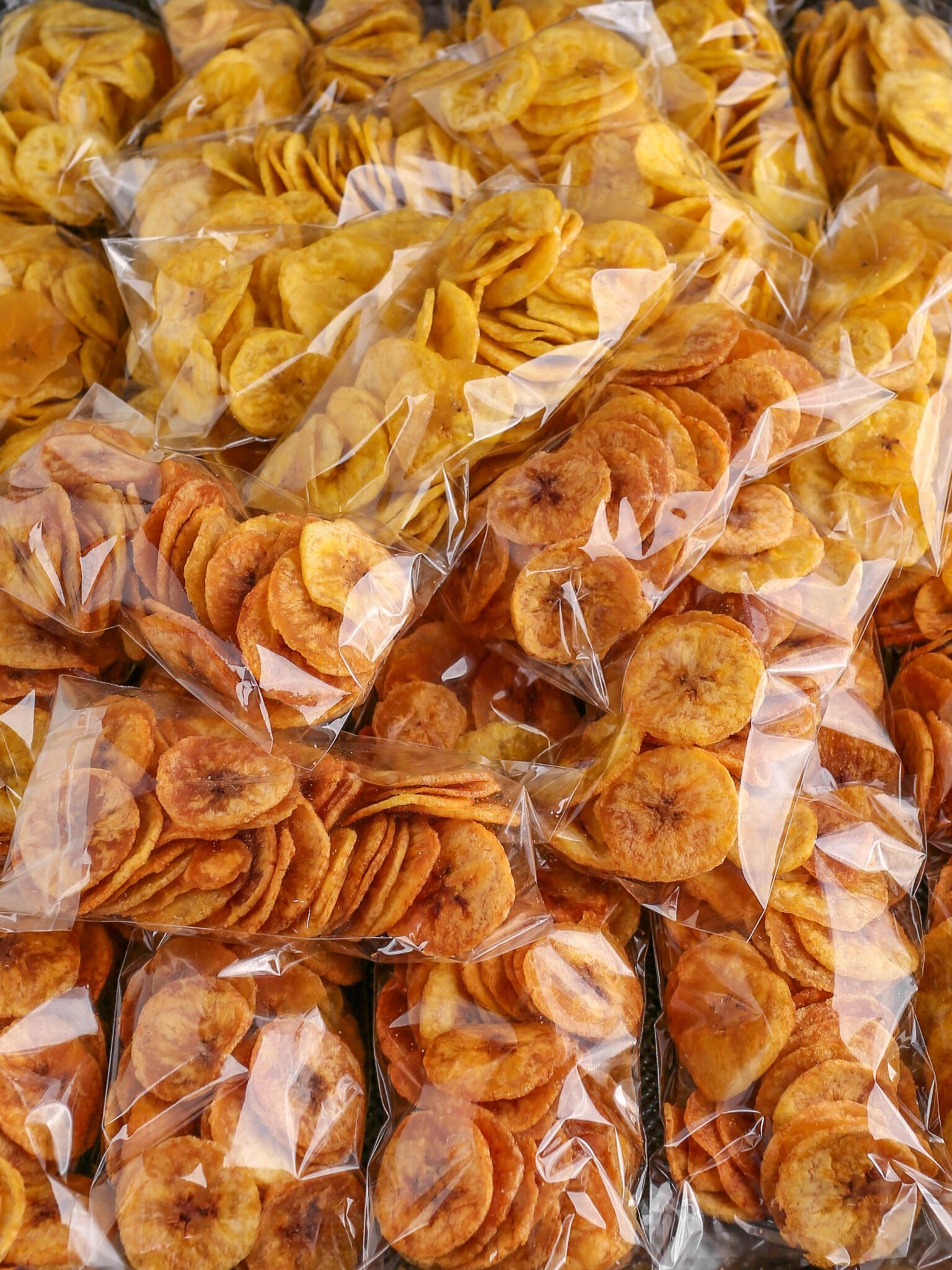 Multiple small clear plastic bags filled with thin, crunchy round plantain chips stacked closely together.