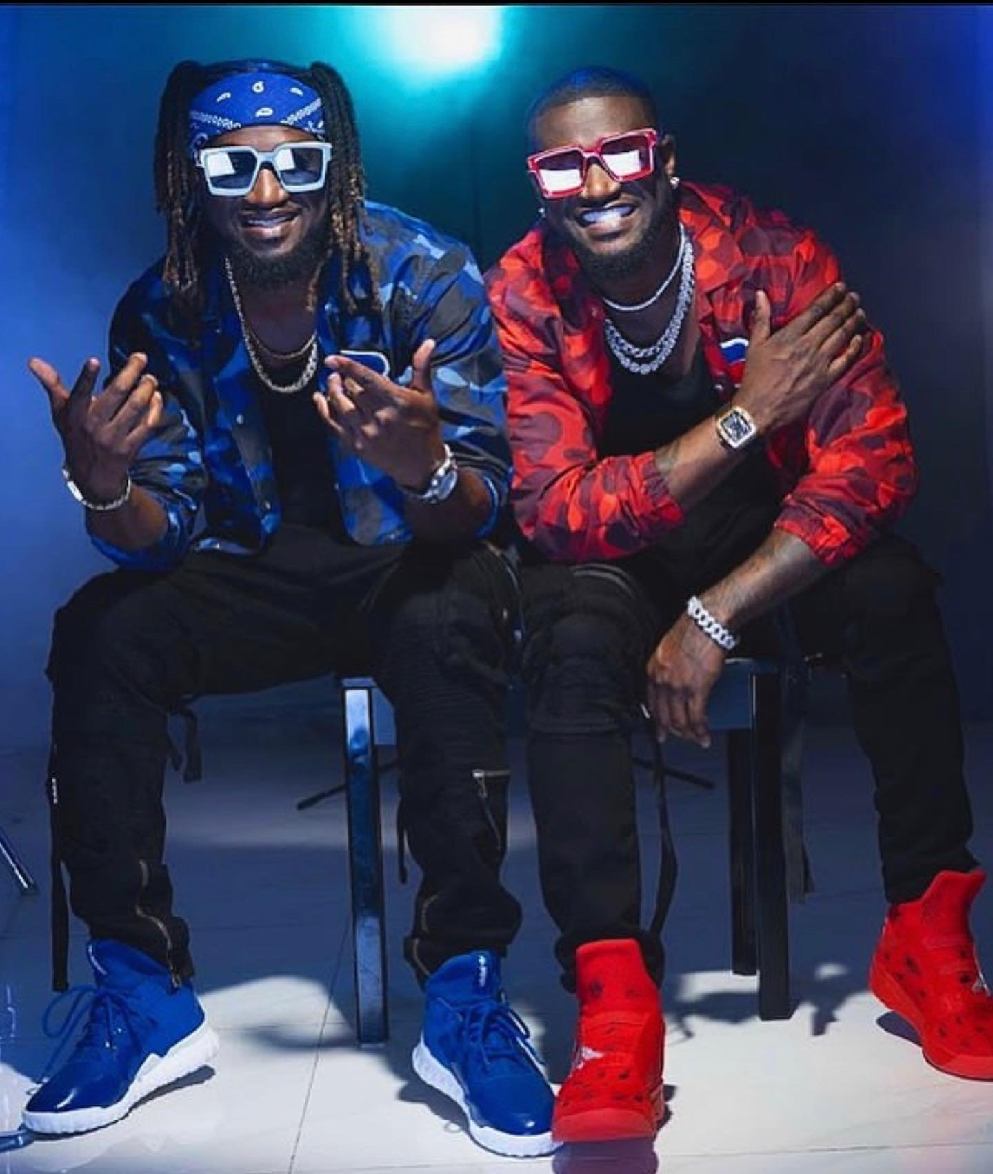Twin brothers Paul and Peter Okoye of P-Square seated together wearing contrasting blue and red camouflage jackets, matching bandanas, sunglasses, and sneakers.