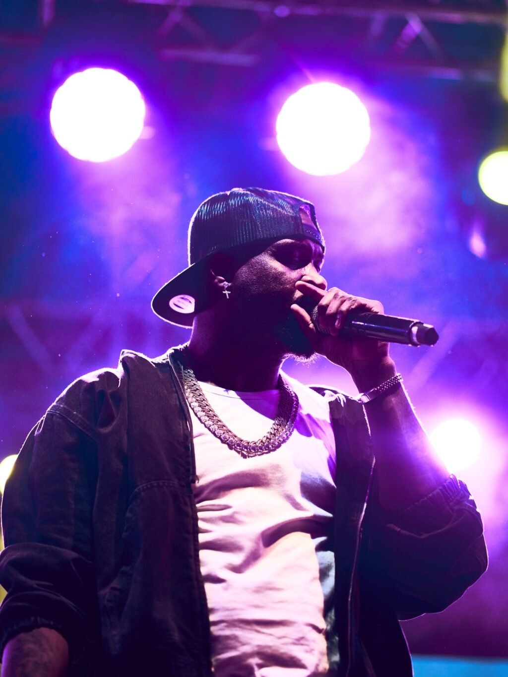 Nigerian singer Mr P performing live on stage under purple spotlighting, holding a microphone while wearing a backward cap, white t-shirt, jacket, and silver chain.