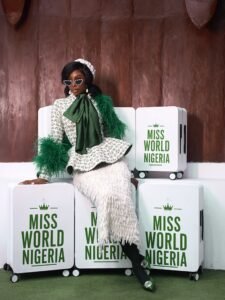 Tamunosoye Karibi-George wearing a green-and-white tweed outfit with green feather trim and sunglasses, sitting on branded Miss World Nigeria suitcases.