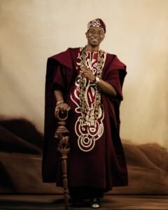 Adedimeji Lateef smiling and pointing forward wearing a burgundy Agbada set with layered coral beads and a carved staff.