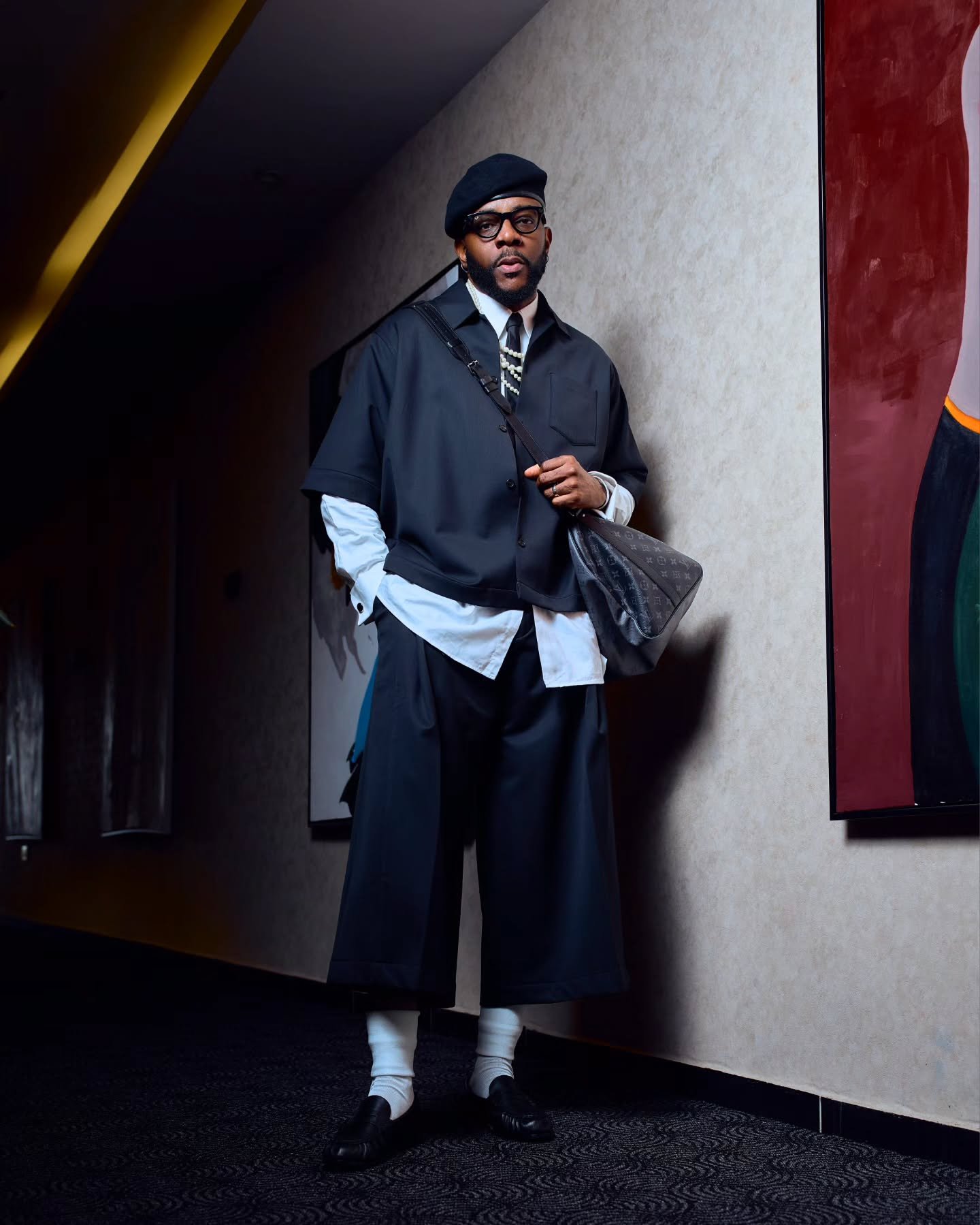 Big Brother Naija Season 11 host Ebuka Obi-Uchendu standing in a black layered short-sleeve shirt over a white button-down, wide-leg cropped trousers, black beret, and loafers.