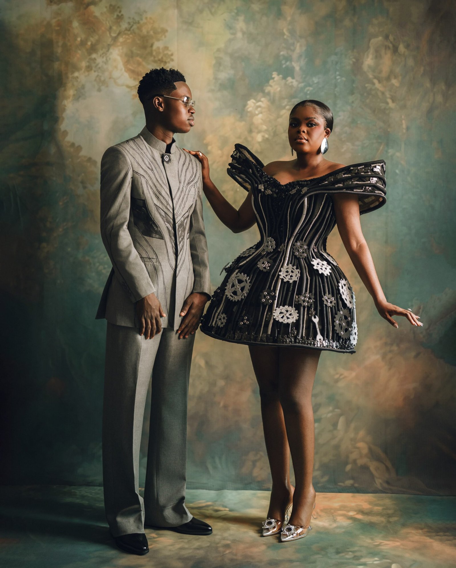 Jarvis in an off-the-shoulder mechanical mini dress standing beside Peller in a tailored grey suit.