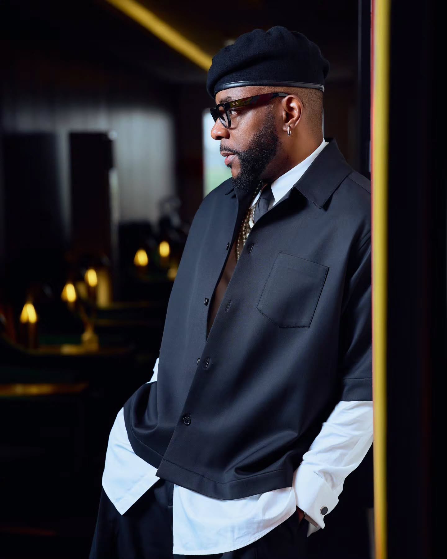 Close-up profile of Ebuka Obi-Uchendu wearing a black beret, black-framed glasses, and a layered collar with a tie and pearl necklace.