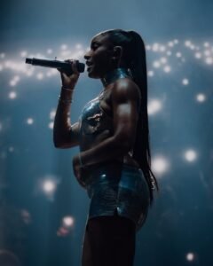 Ayra Starr performing live on stage at All Points East 2026 festival holding a microphone in a metallic silver two-piece outfit with a sleek ponytail.