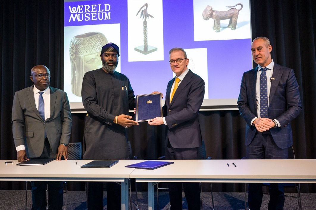 NCMM Director-General Olugbile Holloway holding a signed transfer agreement alongside Dutch Minister Eppo Bruins in front of Benin Bronzes displays at the Wereldmuseum in Leiden.