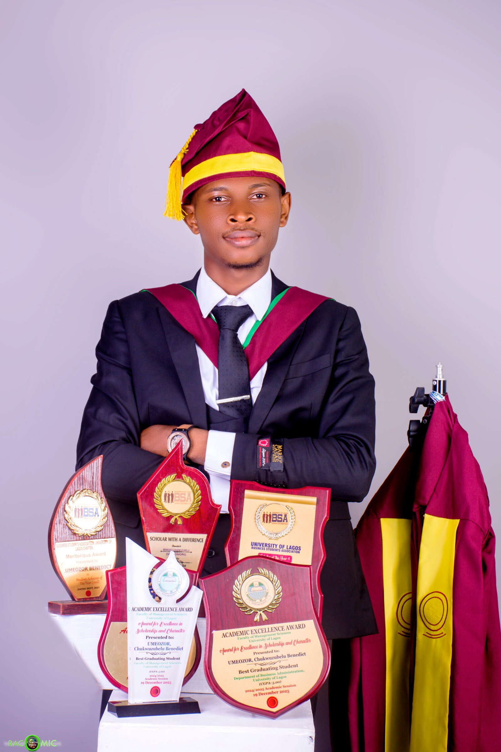 Benedict Umeozor in a three-piece suit standing beside his University of Lagos convocation gown.