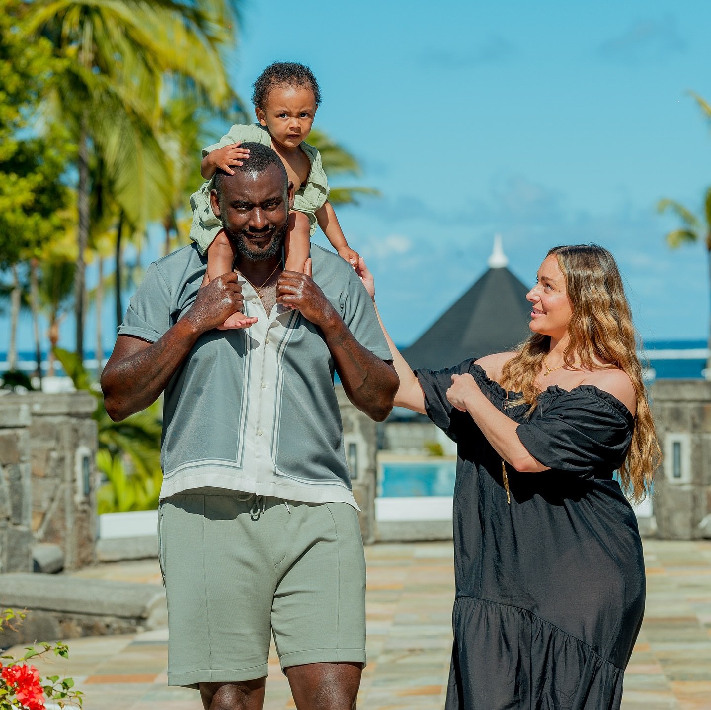 SE Dons captain George Desmond Kamurasi holding his young son on his shoulders alongside his wife Abbey Kamurasi outdoors.