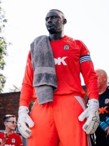 SE Dons captain George Desmond Kamurasi on the touchline wearing a red goalkeeper jersey and captain