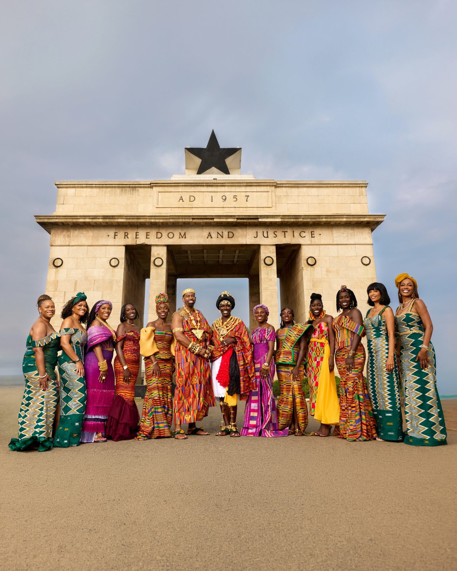 Bozoma Saint John, Keely Watson, and their wedding party posing in matching green and purple Kente outfits in front of the Black Star Monument in Accra.