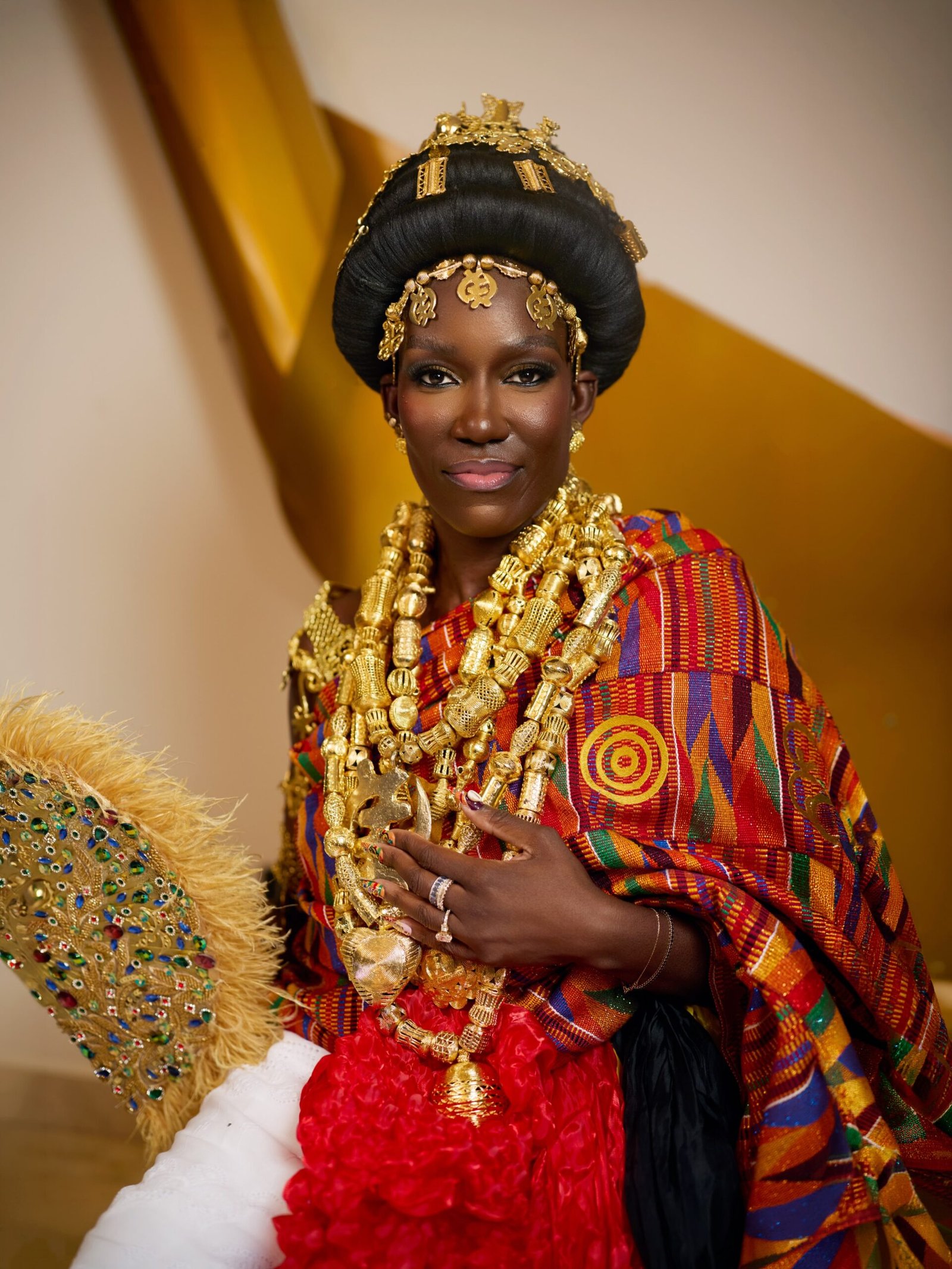 Bozoma Saint John holding a gold feathery jewelled fan in hand-woven Kente cloth and stacked Akan gold jewellery.