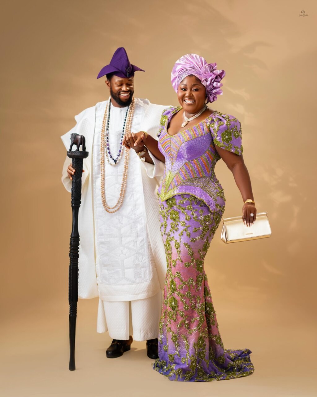 Stan Nze holding hands with Blessing Nze as they smile together in matching white and purple traditional Nigerian outfits.