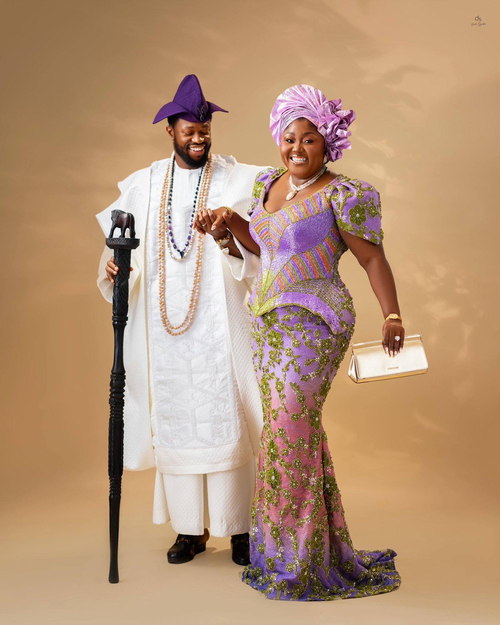 Stan Nze holding hands with Blessing Nze as they smile together in matching white and purple traditional Nigerian outfits.