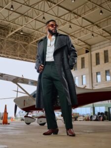Ebuka Obi-Uchendu in a charcoal windowpane trench coat by Deji & Kola layered over a forest-green suit with brown Cuban-heel Chelsea boots.