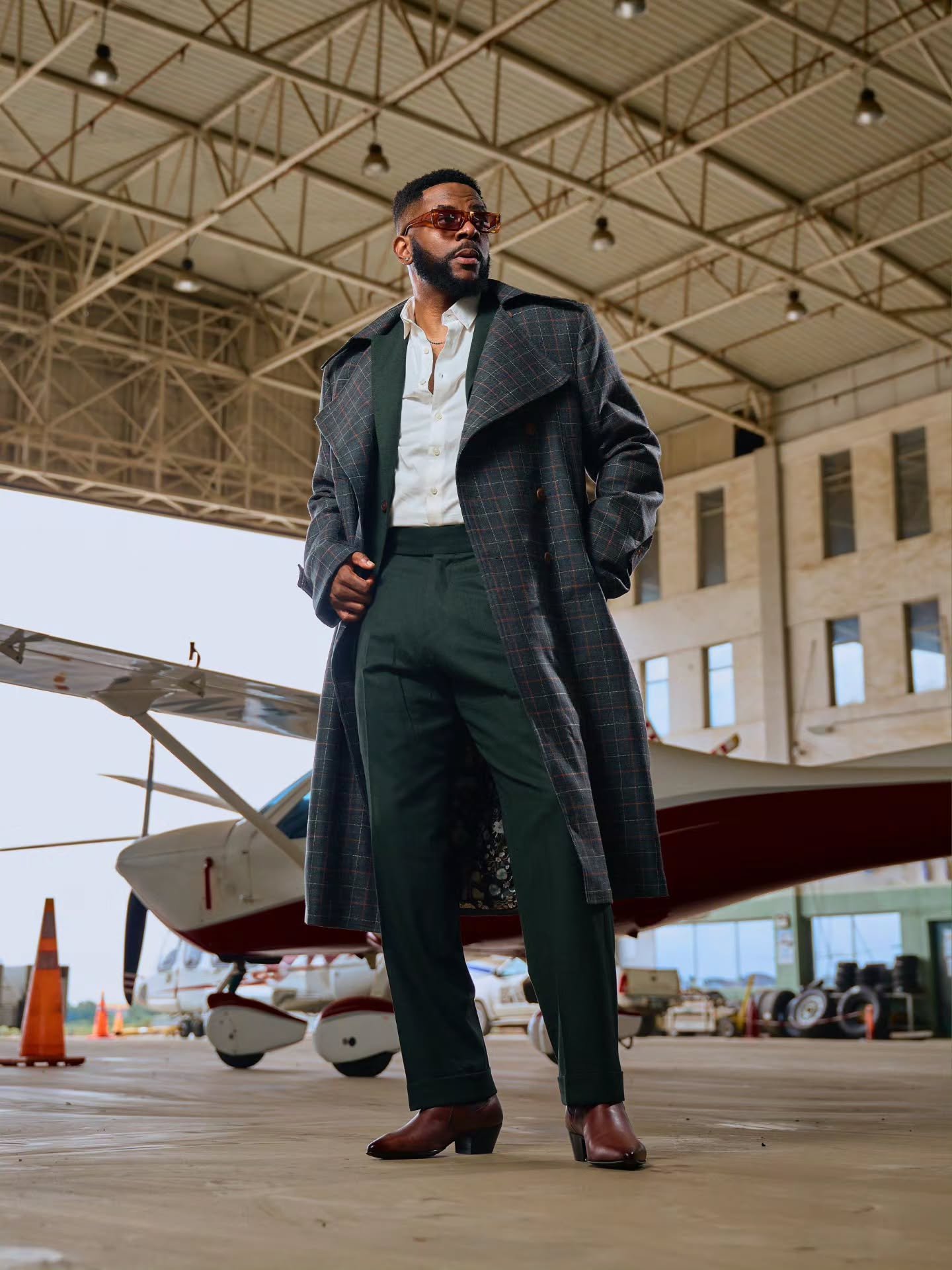 Ebuka Obi-Uchendu in a charcoal windowpane trench coat by Deji & Kola layered over a forest-green suit with brown Cuban-heel Chelsea boots.