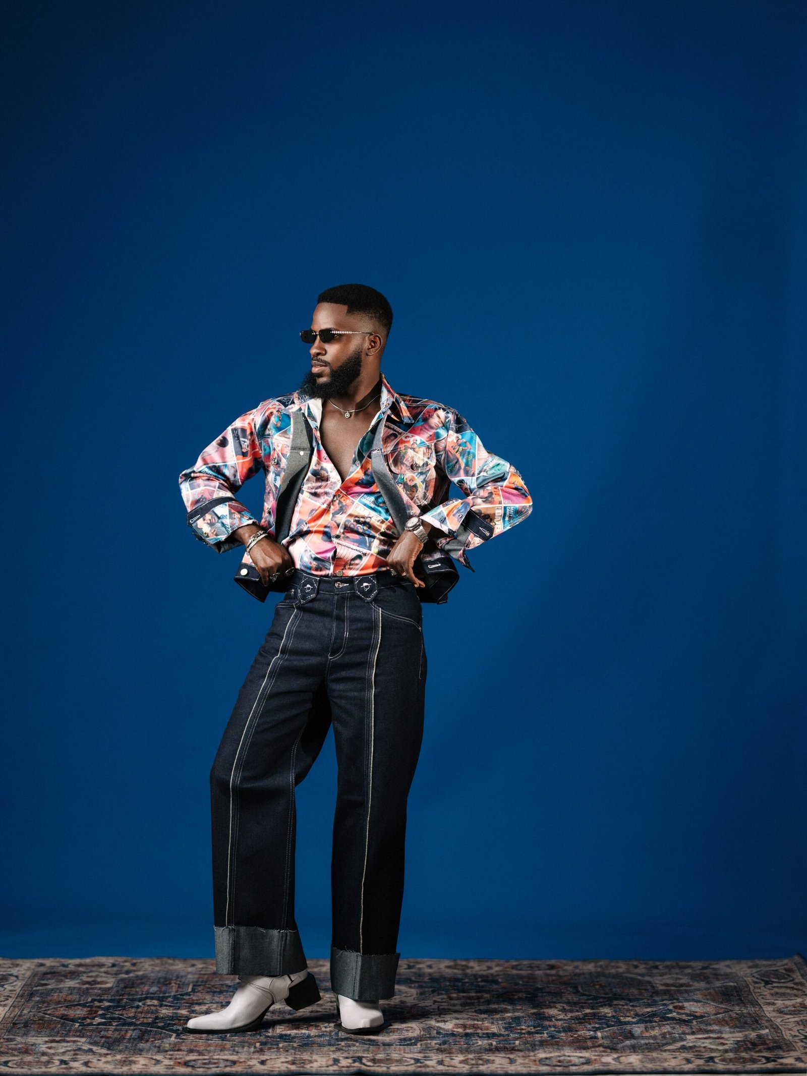 Akin Faminu wearing a Deji & Kola retro magazine print shirt with wide-leg contrast-stitched trousers, sunglasses, and white boots.