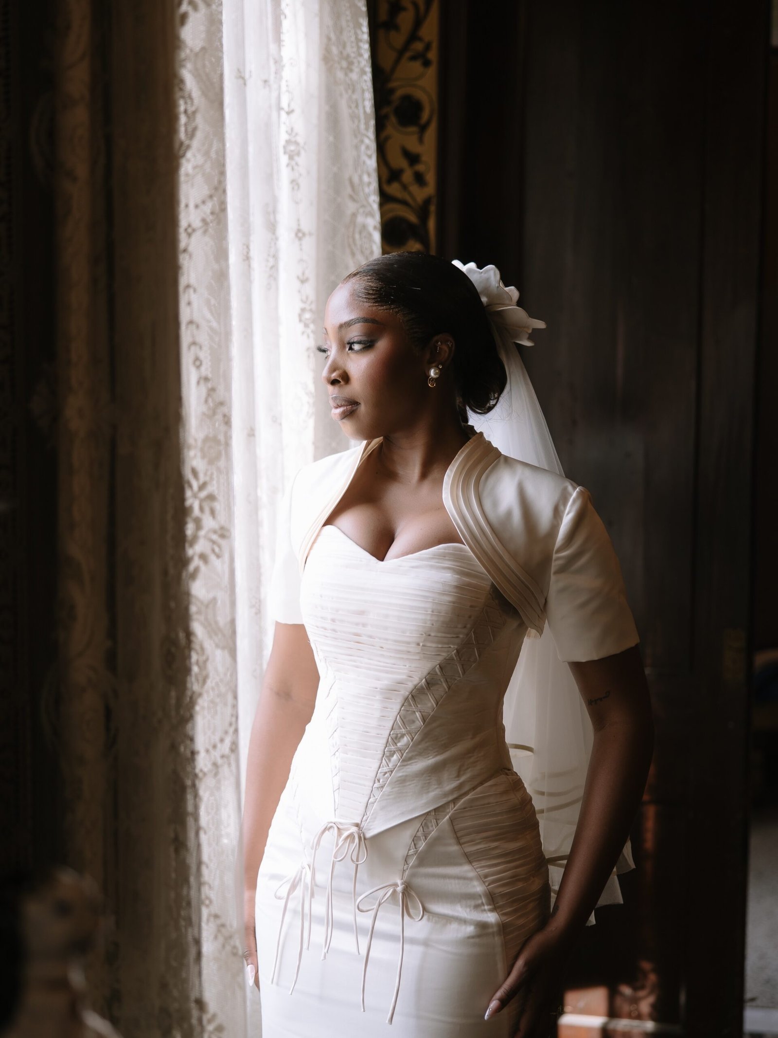 Ella looking out a window wearing a custom Mazelle Bridal gown with a lace-up corset and cropped bolero jacket.