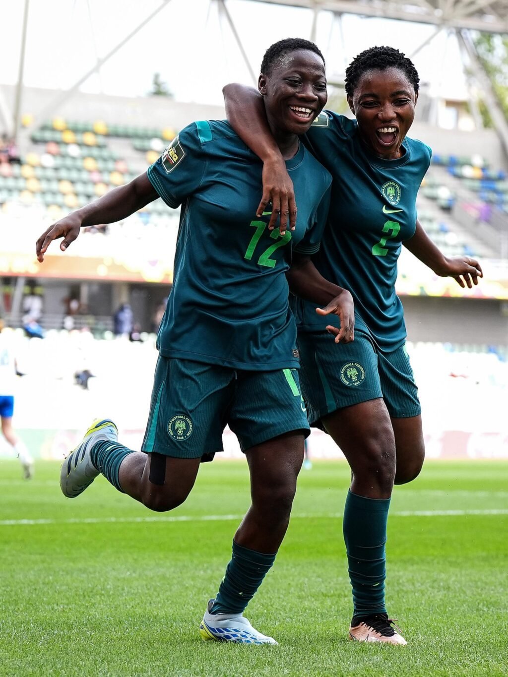 Two Nigeria Falconets players smiling and running side-by-side with arms around each other to celebrate a goal.