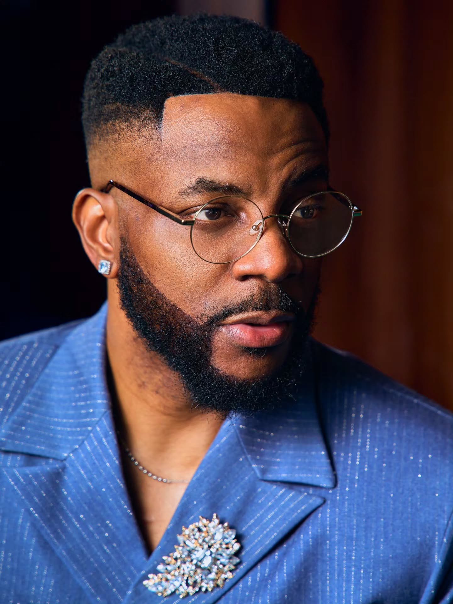 Ebuka Obi-Uchendu wearing round wire-rimmed glasses, diamond stud earrings, and a lapel brooch in custom Atafo menswear.