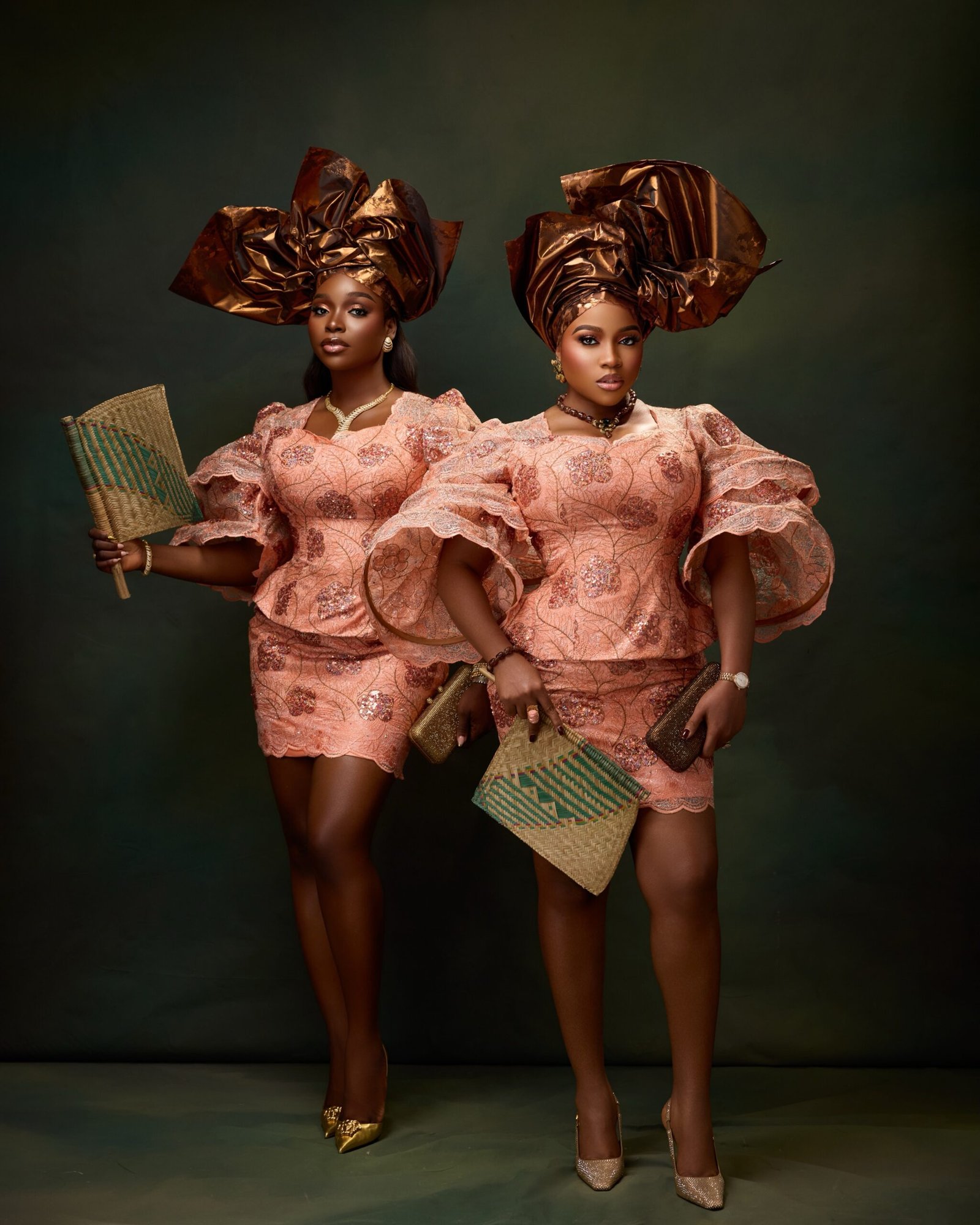Veekee James and Tomike Adeoye posing in knee-length peach lace blouse and skirt sets with bronze-gold structural headties and woven fans