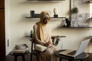 A Muslim woman in a hijab focused on studying in a modern indoor setting with a laptop and notebooks.