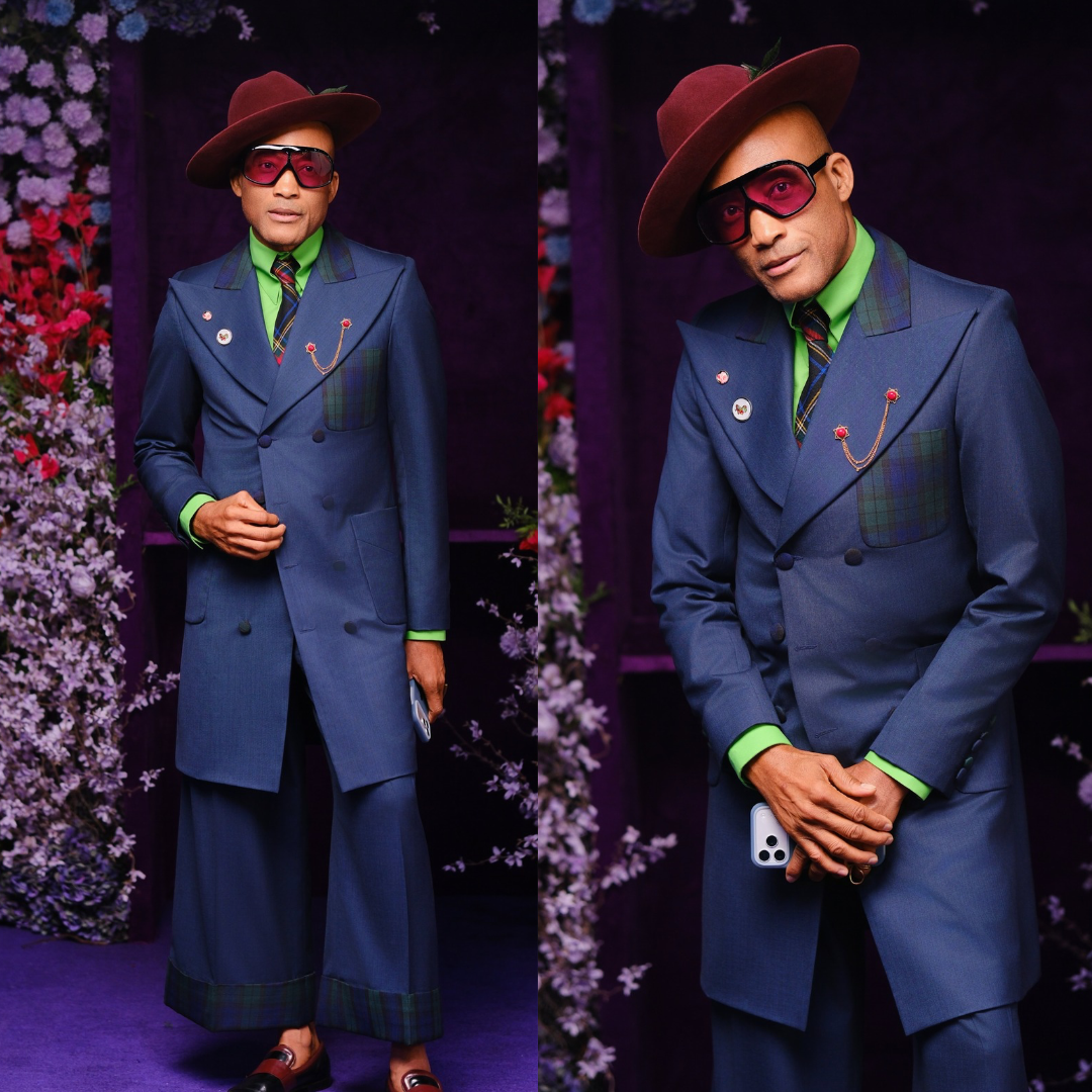 Emmy Collins London wearing a teal blue longline suit jacket with green tartan plaid panels, green shirt, tartan tie, and burgundy fedora.