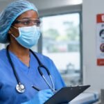 Medical worker in protective scrub cap, safety goggles, face mask, and gloves holding a clipboard near an Ebola prevention poster.