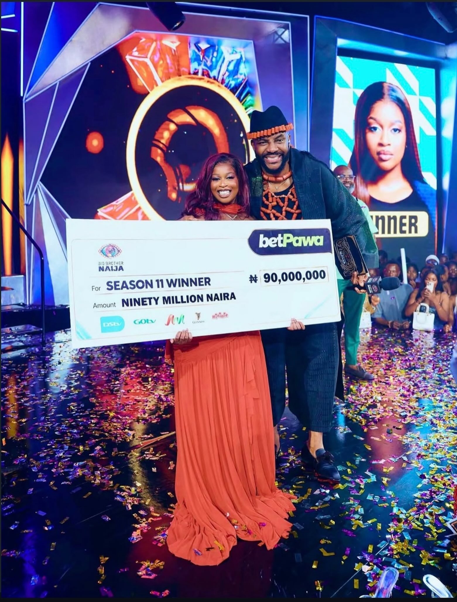 BBNaija Season 11 winner Temi Nkem holding the 90 million Naira cheque alongside host Ebuka Obi-Uchendu on stage.