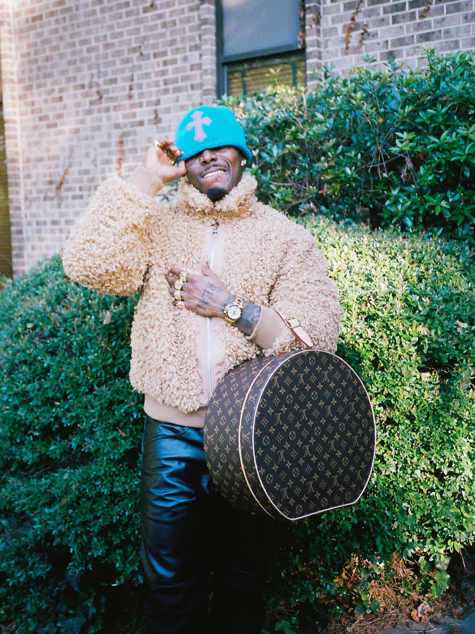 Asake wearing a beige fleece jacket, bright blue beanie, black leather trousers, and holding a round Louis Vuitton monogram hatbox bag.