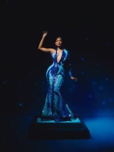 Osas Ighodaro posing on a pedestal in a floor-length mermaid blue mosaic gown by House of Marvee.