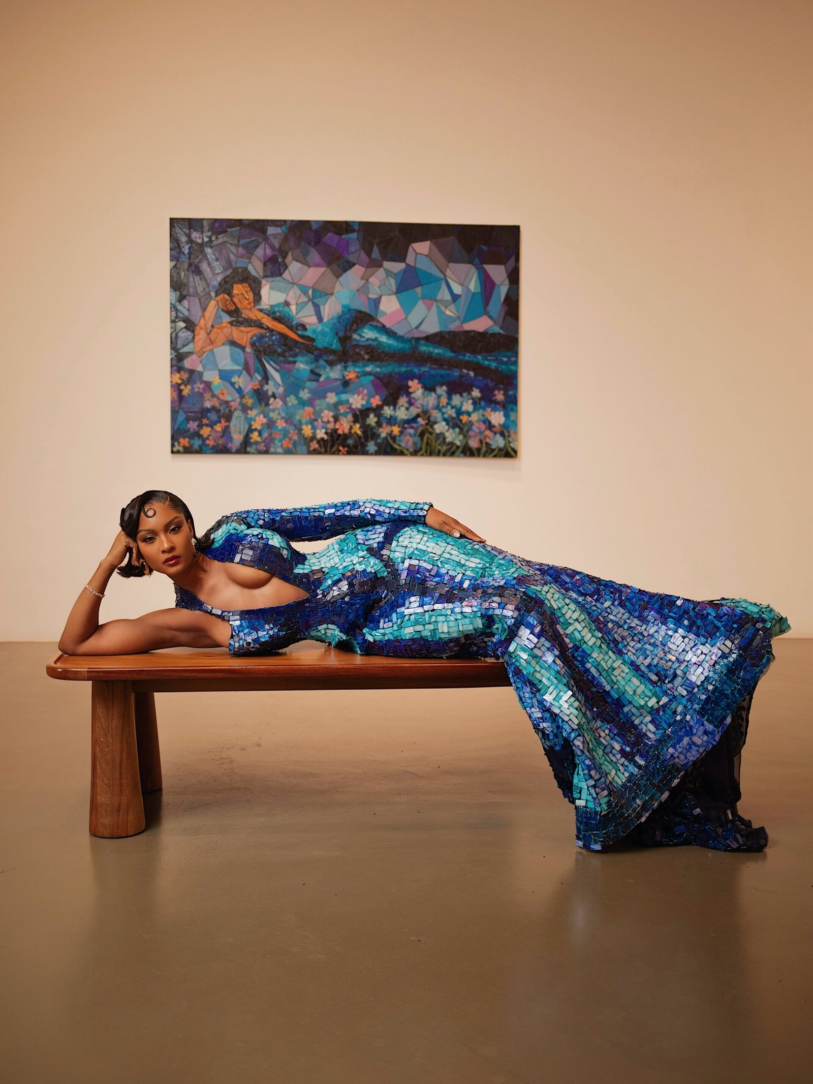 Osas Ighodaro reclining on a wooden bench in a floor-length blue mosaic gown beneath an artwork